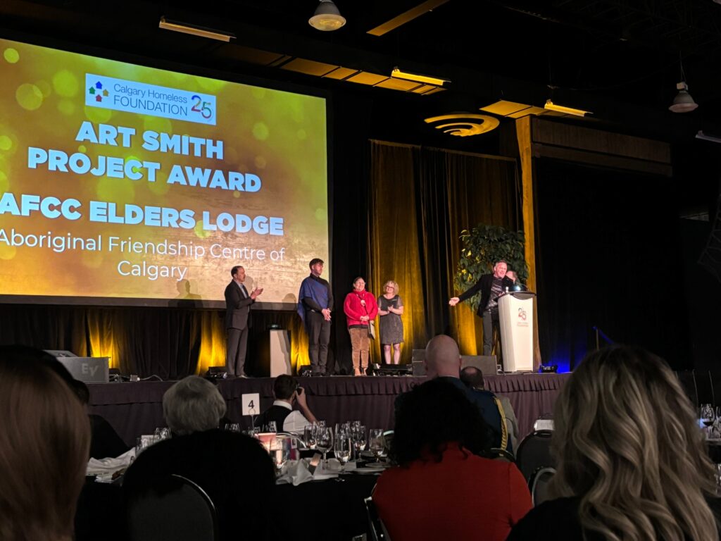 AFCC CEO Shane Gauthier stands at a podium on stage, holding the 2023 Art Smith Project Award for the Elders' Lodge, as he gives an acceptance speech acknowledging the AFCC team during the awards ceremony.
