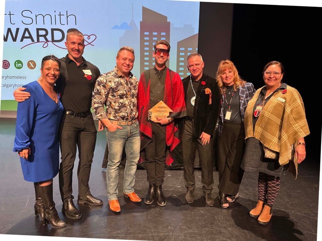 Group photo from the Art Smith Awards 2024, featuring AFCC's Terran Shepard holding his Leadership Award for Collaboration. He is accompanied by fellow AFCC staff and representatives from the Calgary Homeless Foundation, all formally dressed and smiling before an awards banner.