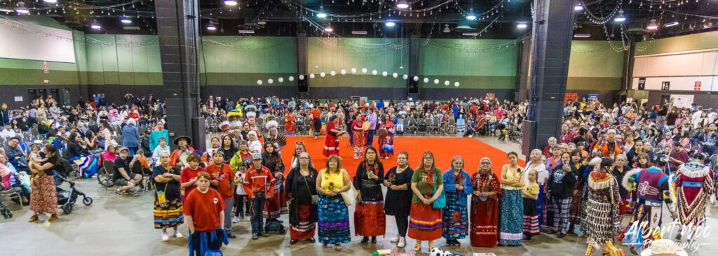 Panoramic photo from the 2nd Annual AFCC MMIWP Red Dress Powwow. A large crowd gathers in a hall to honour victims, with a line of women and Elders in traditional ribbon skirts at the forefront.