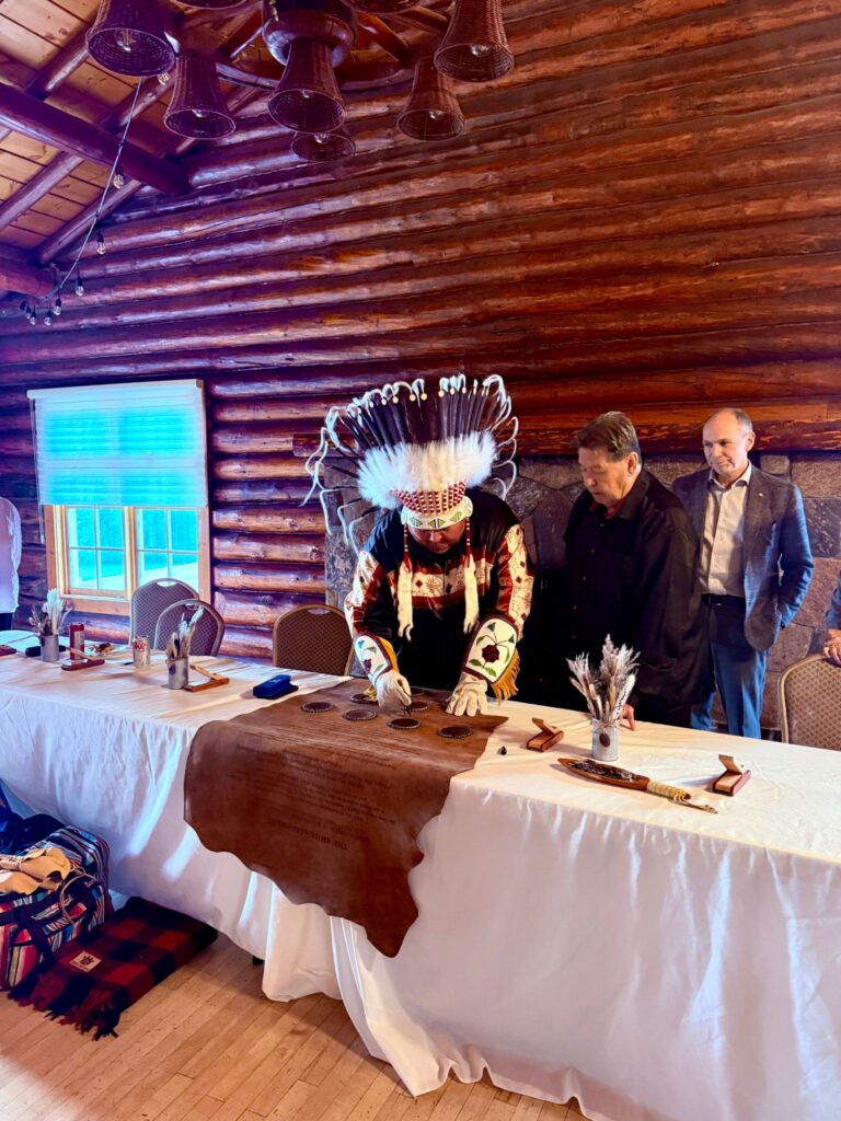 Dr. Tyler White, wearing a traditional feather headdress and regalia, ceremonially signs a hide, marking a new research partnership. Two men in suits stand beside him, observing the signing at a formal event inside a log building.